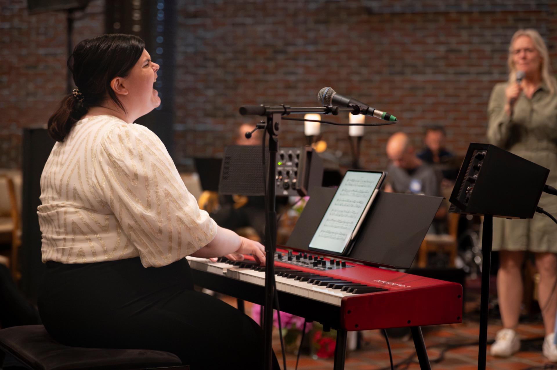 Kvinne spiller piano og synger i konsertsal, mikrofon foran, publikum og bandmedlemmer i bakgrunnen.