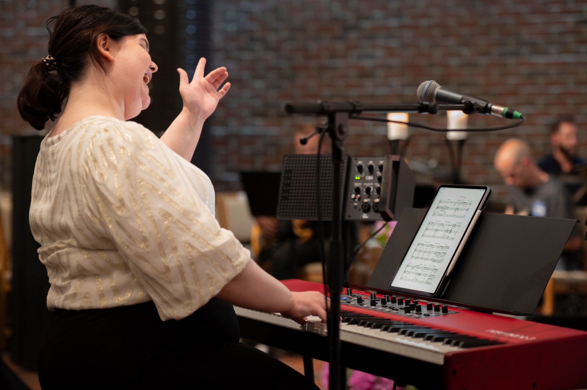 Kvinne ved keyboard i konsertsal, dirigerer kor med en hånd og spiller piano med den andre